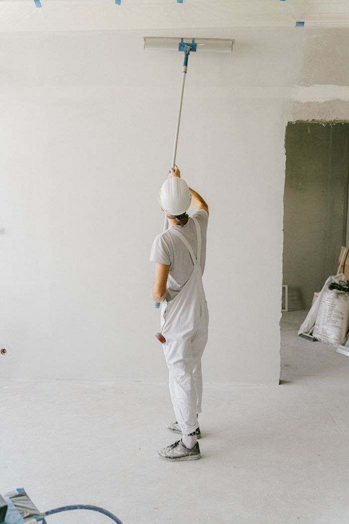 services-02 Back view of a painter in overalls and hard hat painting an interior wall with a roller.